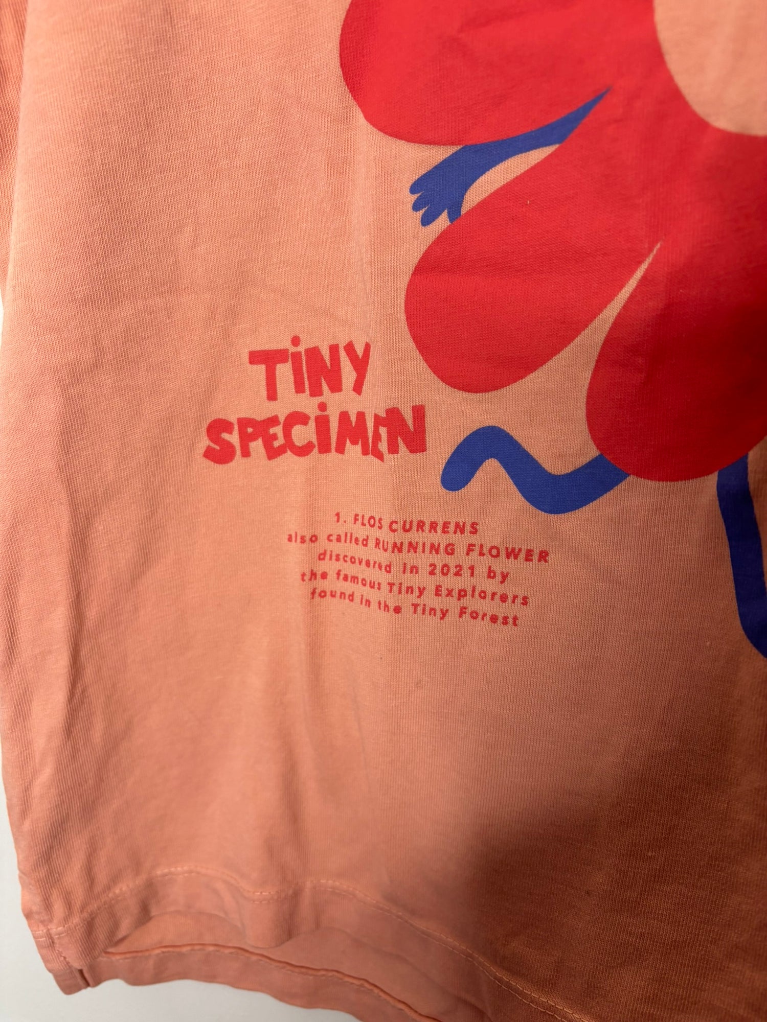 Peach T-shirt featuring a red flower with a smiling face and 'Tiny Specimen' text, hanging on a black hanger.