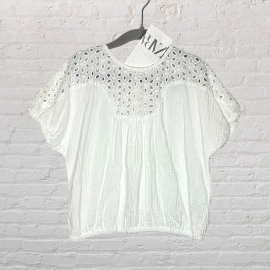 White Zara blouse with eyelet details on the upper part and loose sleeves, hanging against a brick wall.