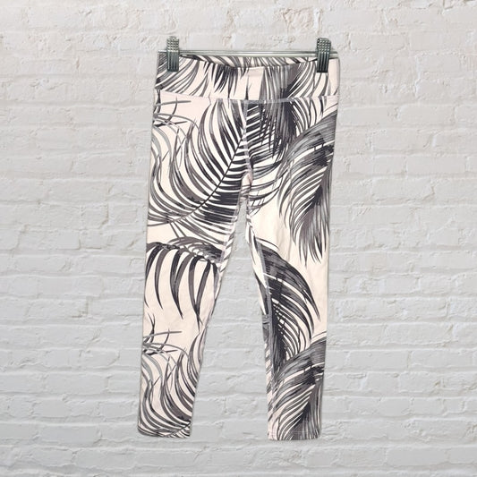 White leggings with a black palm leaf print hanging on a hanger against a white brick wall.
