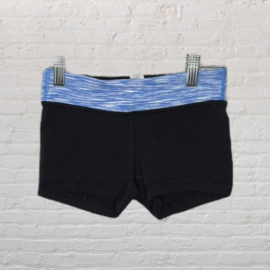 Black gymnastics shorts with a blue patterned waistband hanging on clips against a white brick background.