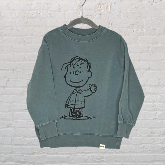 Green sweatshirt with a black line drawing of a Peanuts character on the front, displayed on a hanger against a white brick wall.