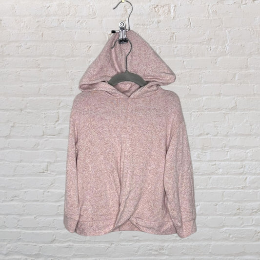 A pink twist front hooded sweater hanging on a gray hanger against a brick wall.