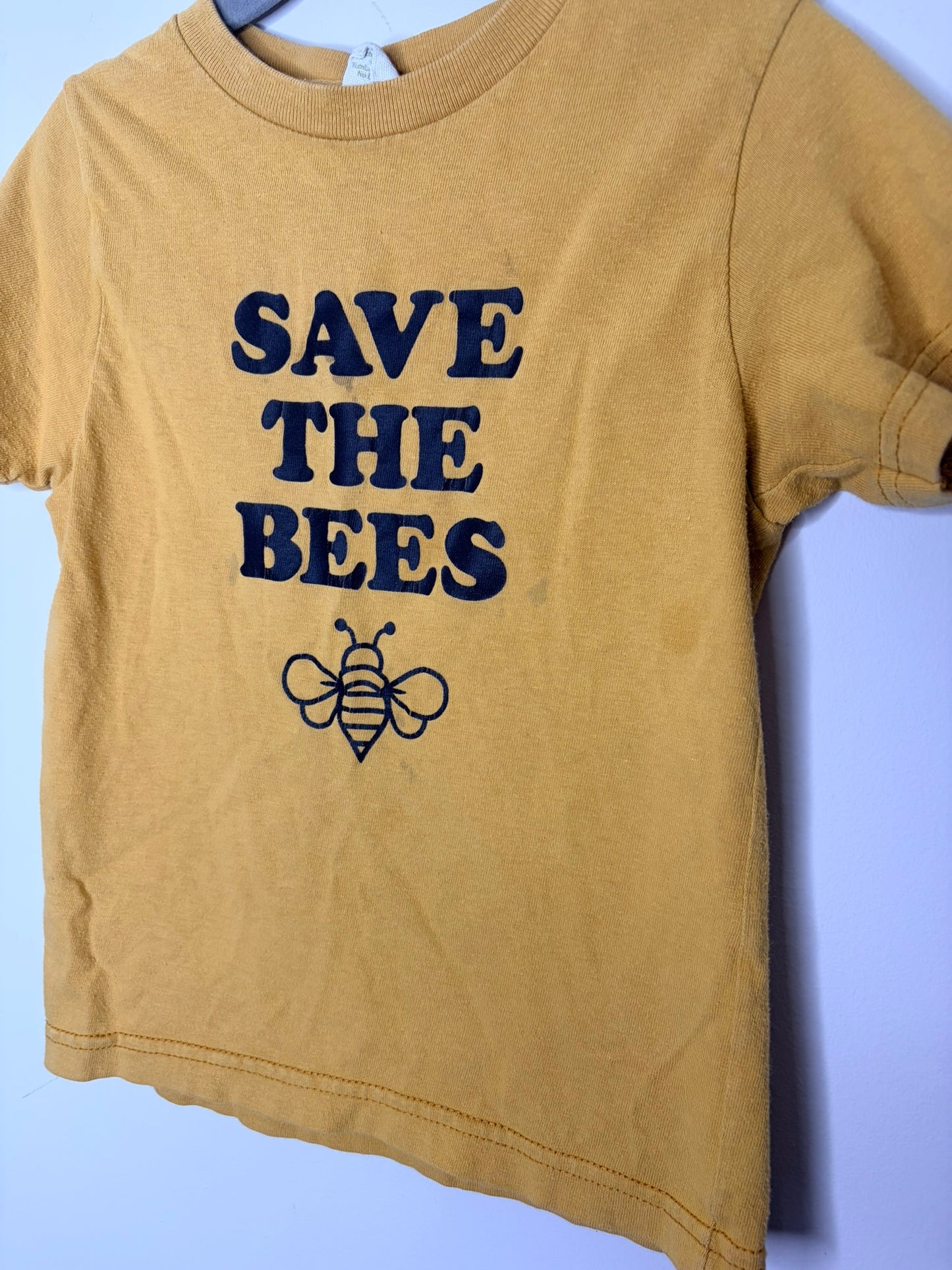 Mustard yellow T-shirt with 'Save The Bees' text and bee graphic, hanging on a hanger.