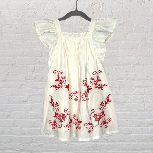 White dress with red floral embroidery and flutter sleeves, hanging on a black hanger against a brick wall.