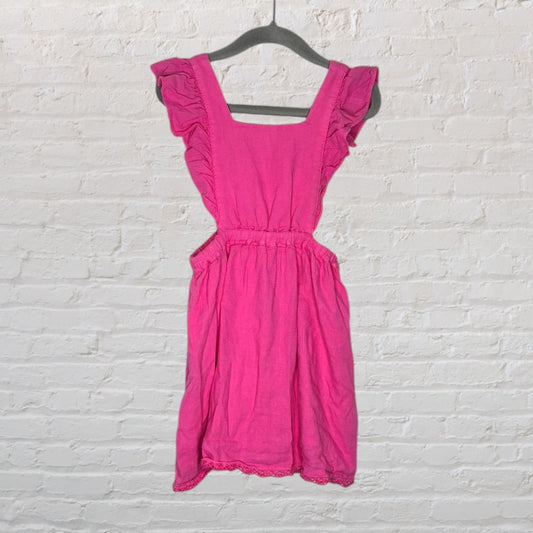 Pink dress with ruffle shoulders and cutout sides, hanging on a black hanger against a white brick background.
