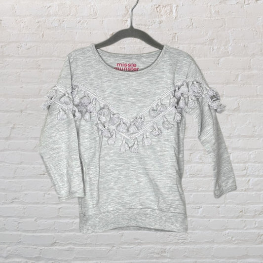 Light grey sweater with fringe detail across the chest and sleeves by Missie Munster, hanging on a hanger.