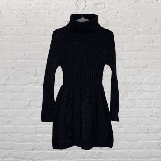 Black cable knit turtleneck sweater dress with long sleeves, hanging on a hanger against a white brick wall.