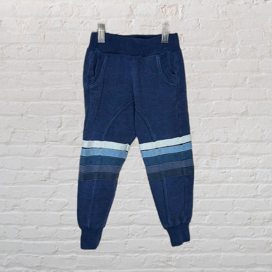 Navy blue jogger pants for children with light blue horizontal stripes, displayed on a hanger against a white brick wall.