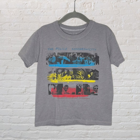 Grey T-shirt featuring 'The Police Synchronicity' multicolor graphic design displayed on a hanger against a white brick wall.