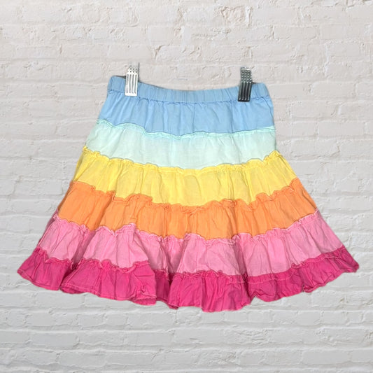 A vibrant rainbow tiered skirt with layers of pink, orange, yellow, and blue ruffles hanging against a white brick background.