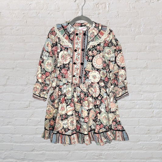 Women's vintage-style dress with a multicolour floral patchwork pattern, featuring long sleeves and decorative buttons, hanging against a white brick wall.