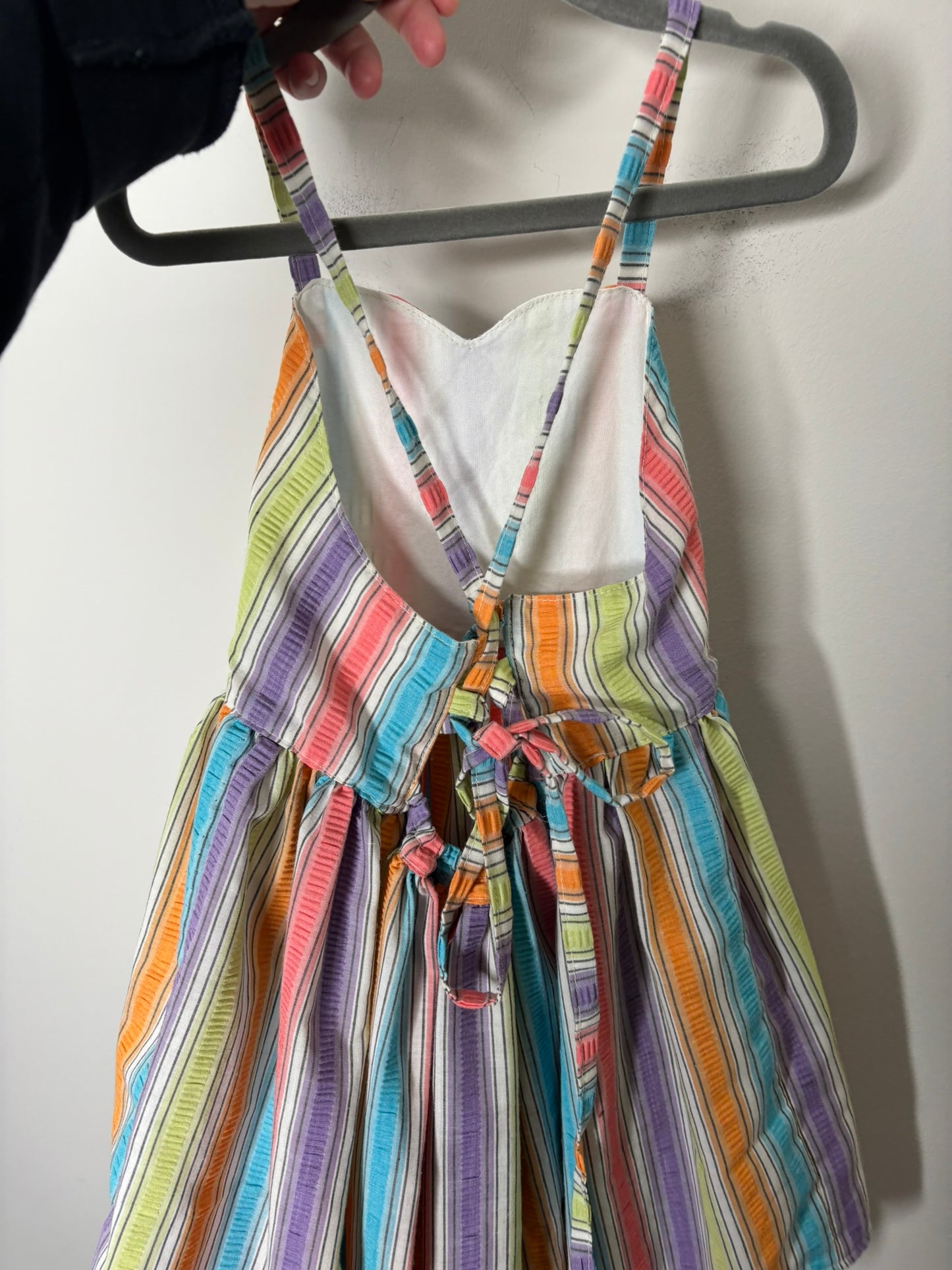 A vibrant rainbow-striped sundress with spaghetti straps, hanging on a black hanger against a white brick wall.