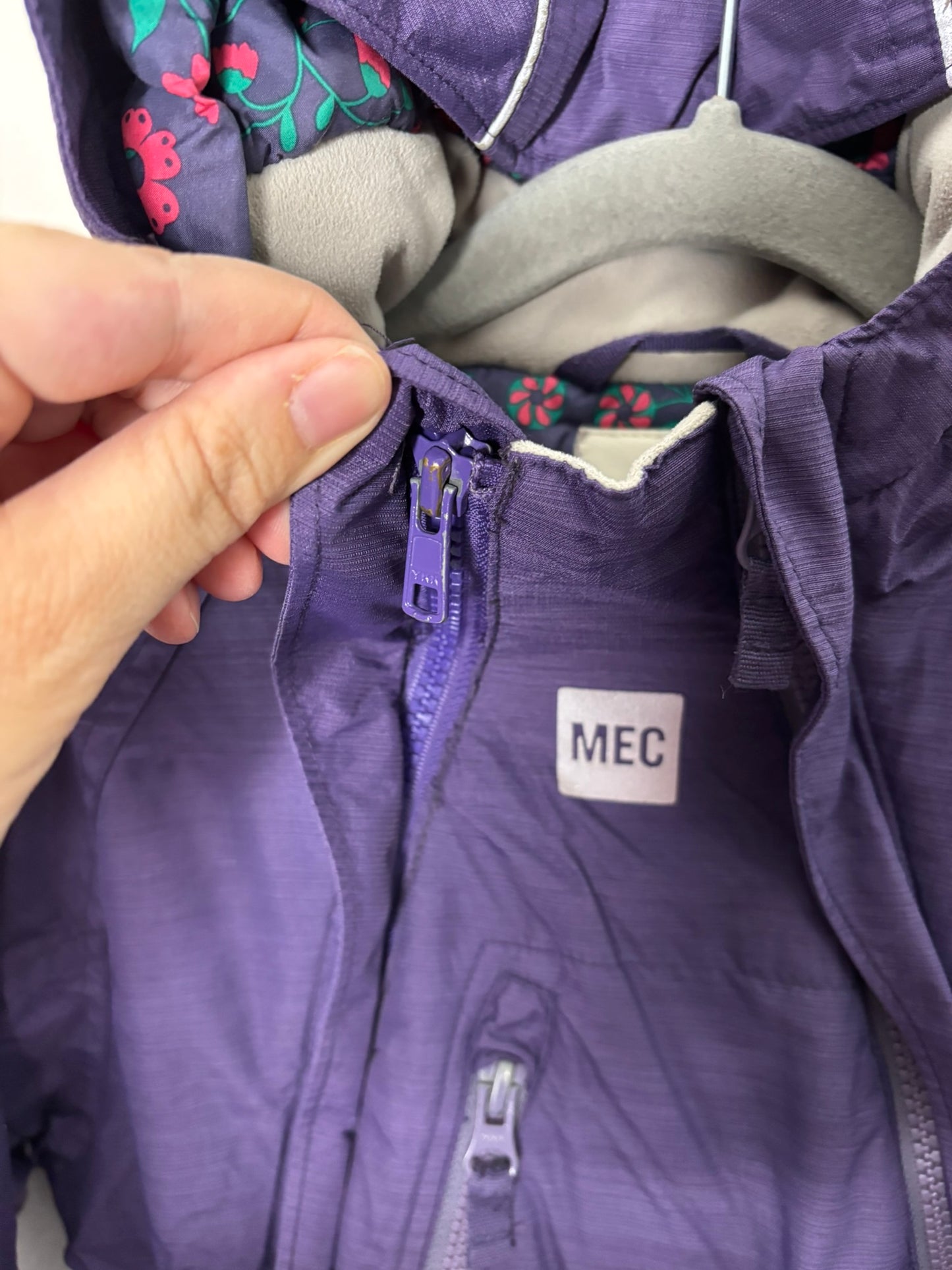 Purple insulated kids' snowsuit with hood and zip closure, hanging against a white brick wall.