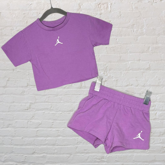 Purple athletic outfit including a crop top and shorts with Jordan logo on a hanger against a white brick background.