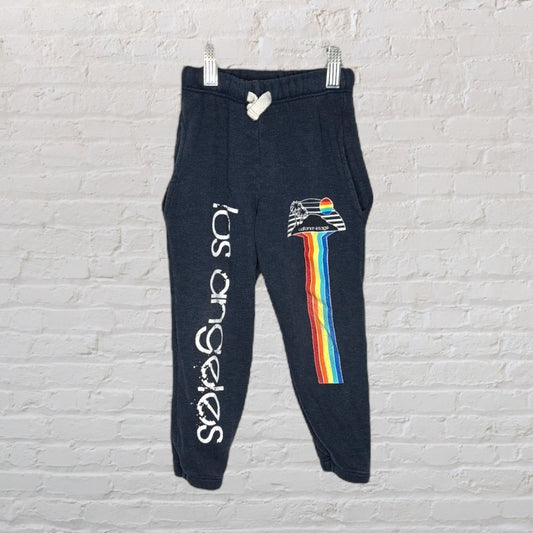 Dark grey sweatpants with Los Angeles text and rainbow graphic on a brick wall background.