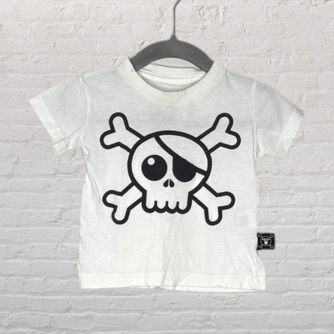 White T-shirt with a black skull and crossbones graphic, hanging on a hanger against a brick wall.