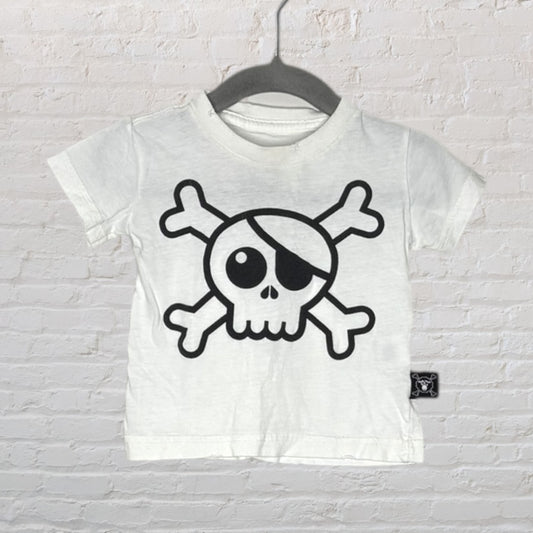 White T-shirt with a black skull and crossbones graphic, hanging on a hanger against a brick wall.