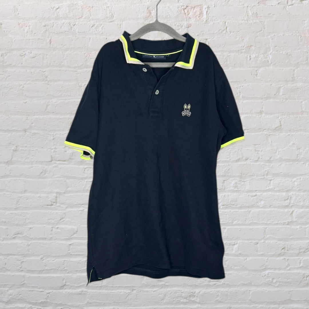 Navy Psycho Bunny polo shirt with neon yellow trim hanging on a grey hanger against a white brick wall.