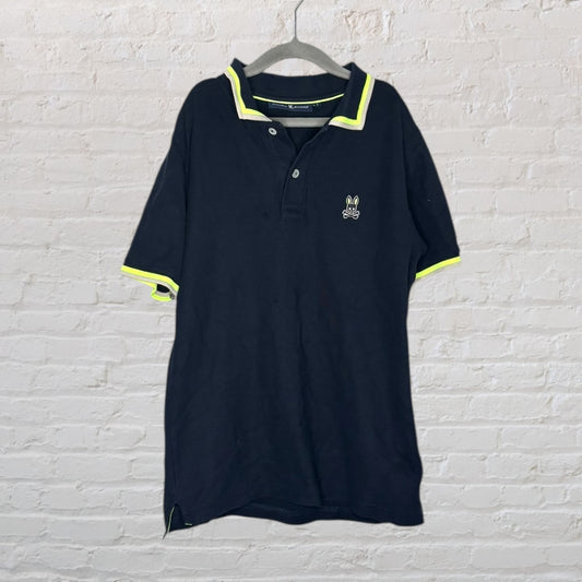 Navy Psycho Bunny polo shirt with neon yellow trim hanging on a grey hanger against a white brick wall.