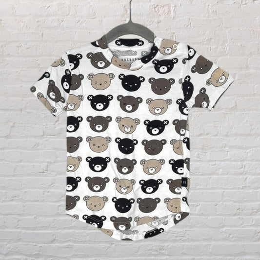 White T-shirt with black and brown cartoon bear prints by Huxbaby hanging against a brick wall.