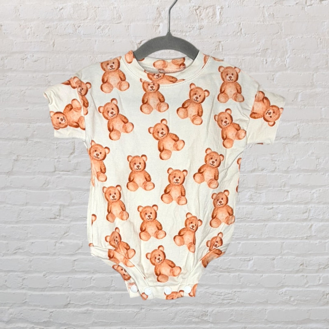 Cream baby onesie with a teddy bear print pattern hanging on a grey hanger against a white brick background.