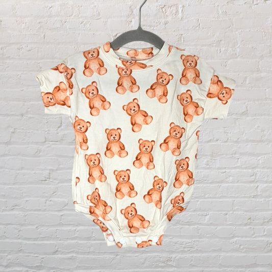 Cream baby onesie with a teddy bear print pattern hanging on a grey hanger against a white brick background.