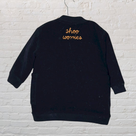 Black jacket with 'Shoo Worries' embroidery on the back, displayed against a white brick wall.