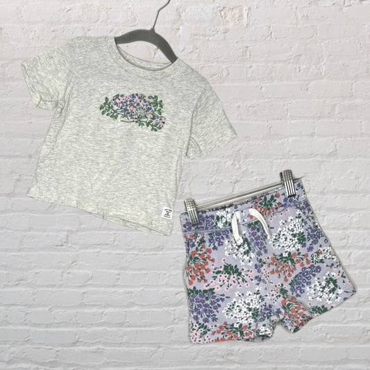 A light grey T-shirt with a floral print and matching multicolor floral shorts hanging against a white brick background.