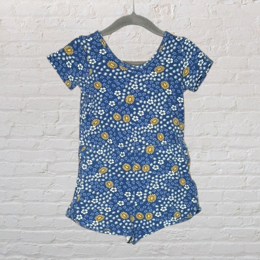 Blue women's romper with a floral pattern in yellow and white, shown on a hanger against a brick wall.