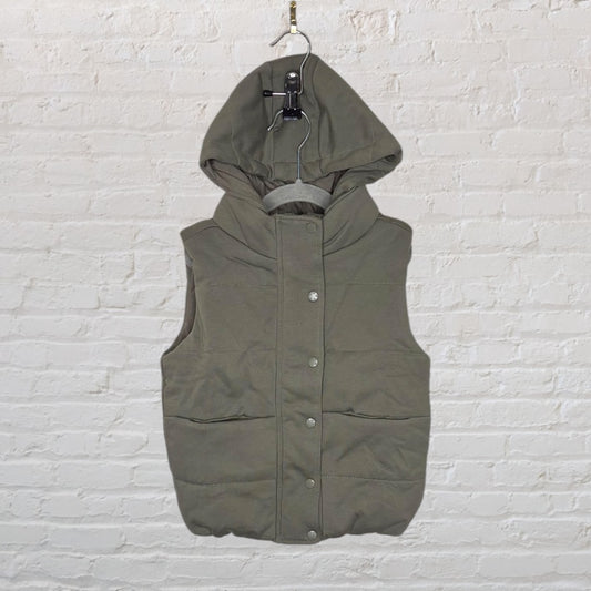 Olive green hooded puffer vest with button closure, hanging in front of a white brick wall.