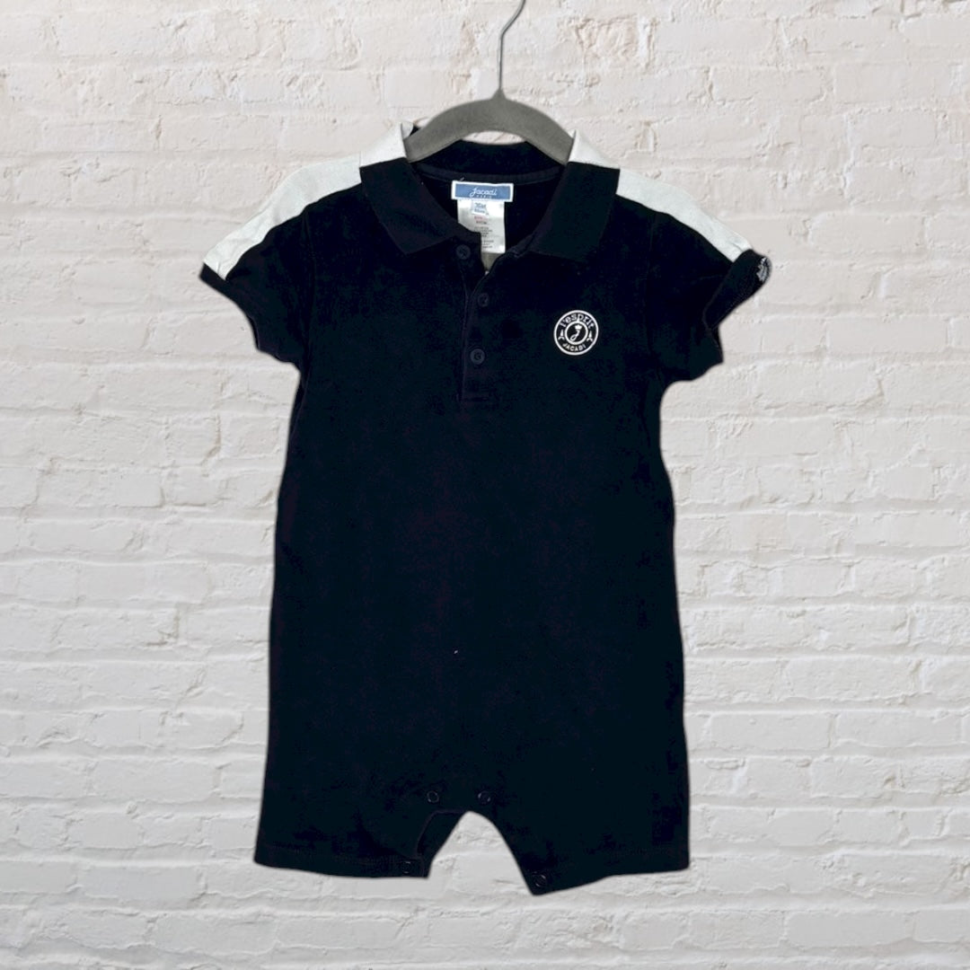Black polo-style romper with white sleeves and a small embroidered logo, hanging on a hanger against a brick wall.