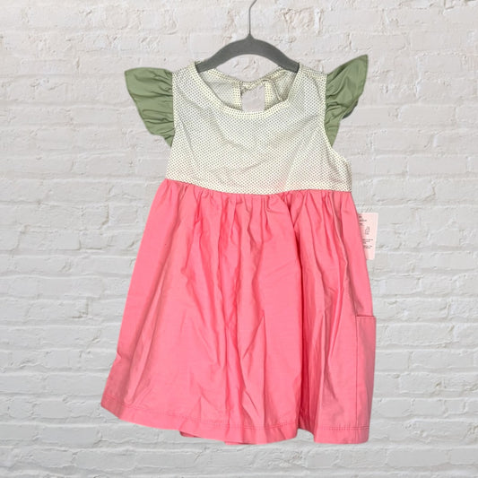 Pink dress with a white polka dot bodice and green ruffle sleeves, hanging on a hanger against a white brick wall.