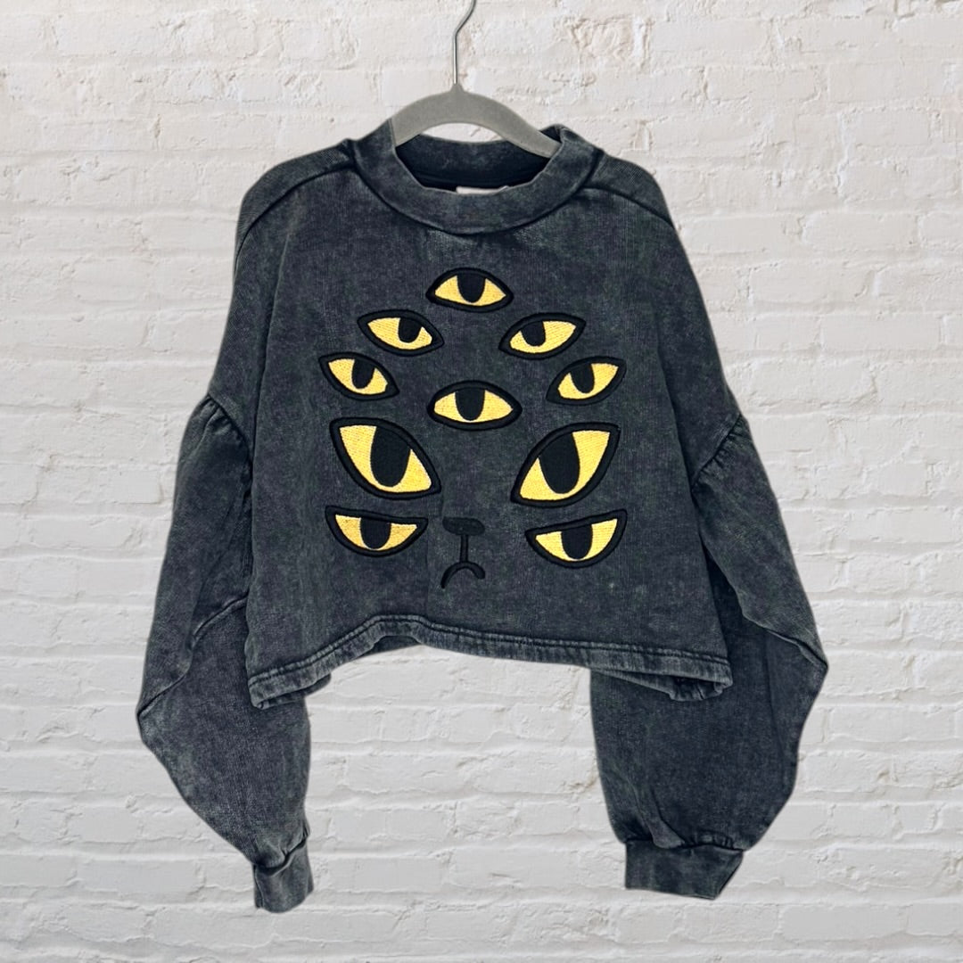 Grey cropped sweatshirt with multiple yellow cat eyes graphic design, displayed on a hanger.