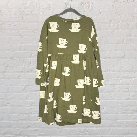 Olive green dress with cream-colored tea cup pattern, long sleeves, hanging on a hanger against a white brick wall.