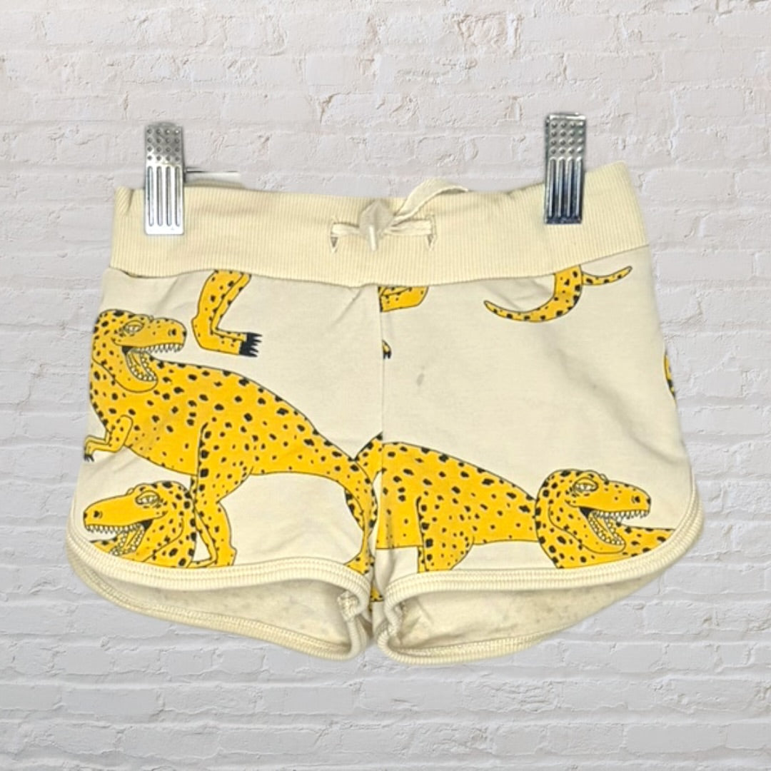 Beige shorts with yellow dinosaur print, displayed on a white brick background with clips.