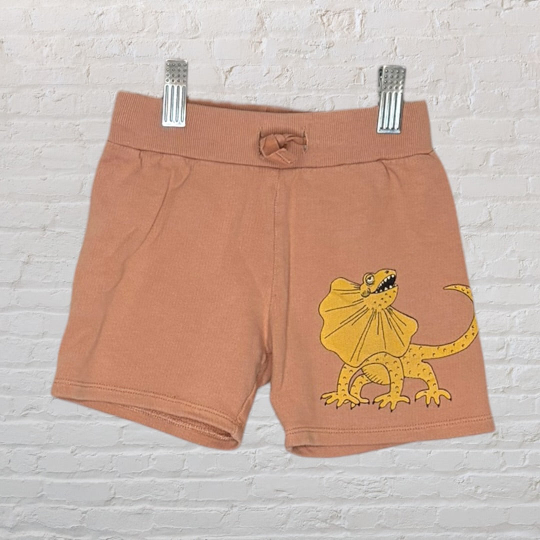 Brown kids shorts with a yellow lizard graphic, displayed on a hanger against a white brick wall.