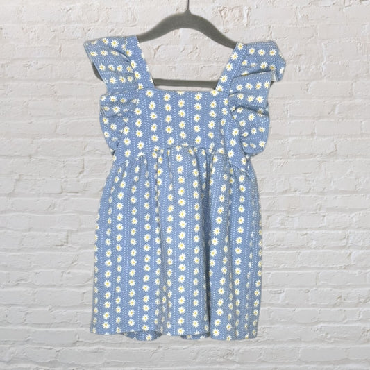 A blue dress with white daisy print and flutter sleeves, hanging on a hanger against a white brick wall.