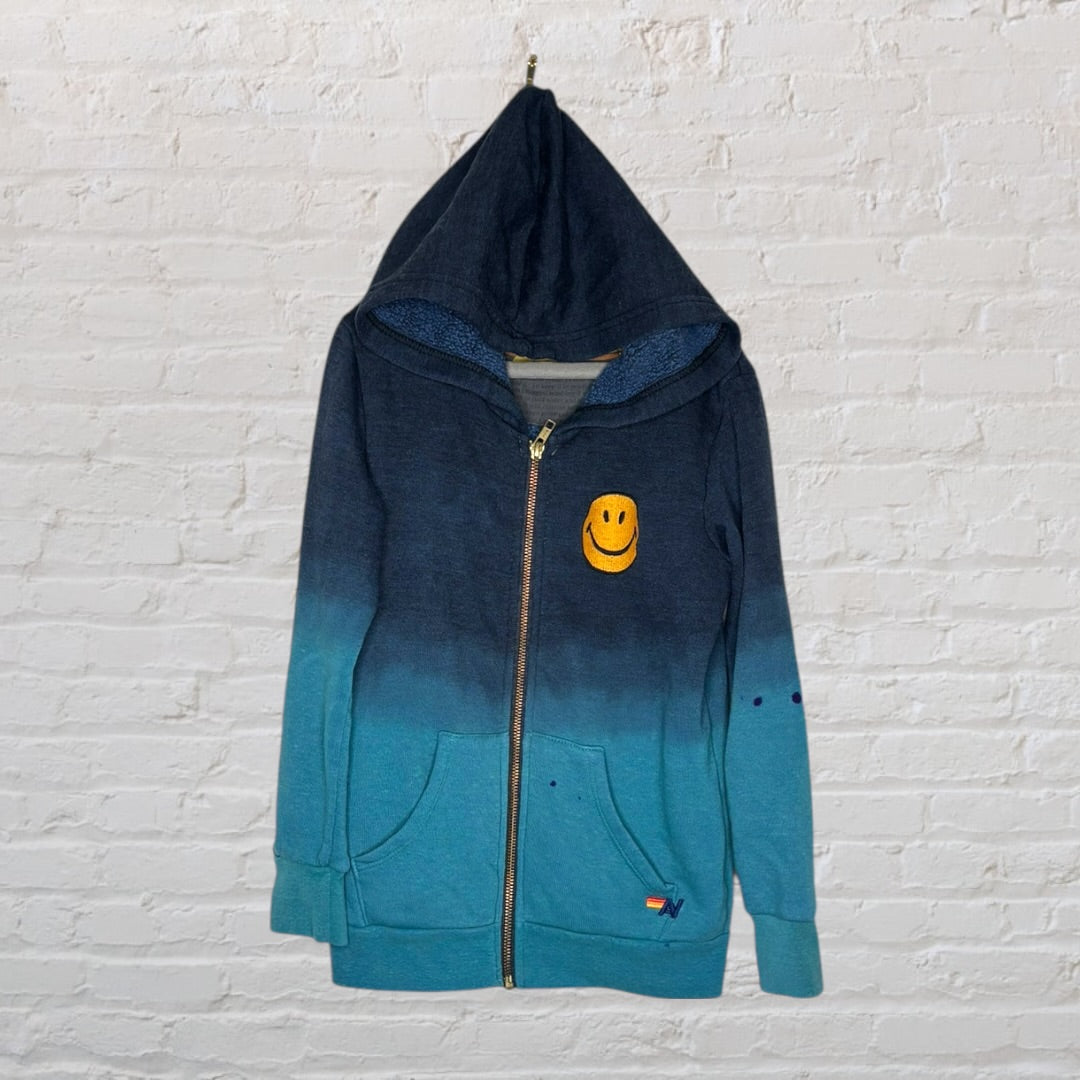 Navy and teal gradient zip-up hoodie with a smiley face patch and pockets, hanging on a white brick wall.