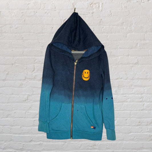 Navy and teal gradient zip-up hoodie with a smiley face patch and pockets, hanging on a white brick wall.