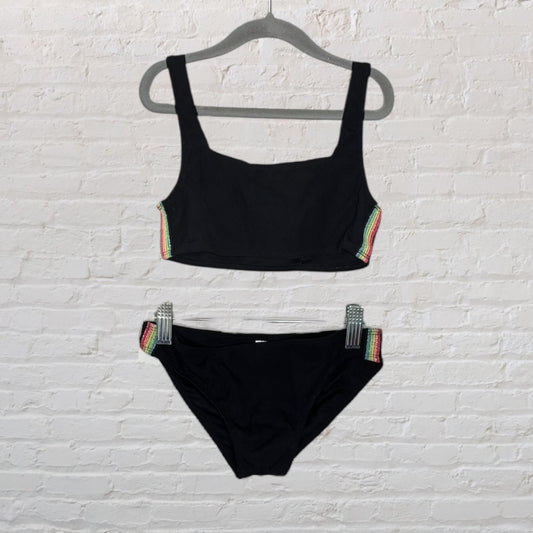 Black two-piece swimsuit with rainbow trim on sides, top and bottom displayed on hangers against a white brick wall.