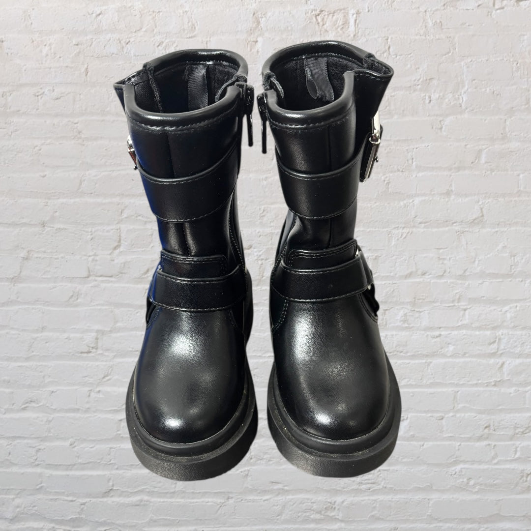 Pair of chunky black boots with silver buckle accents and a thick sole against a white brick background.