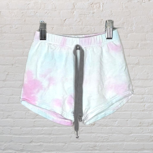 Pastel tie-dye shorts with a grey drawstring, hung on a hanger against a white brick wall.
