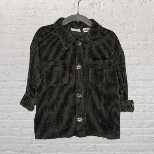 Dark green corduroy button-up jacket with pockets and folded sleeves, hanging on a hanger against a brick wall.