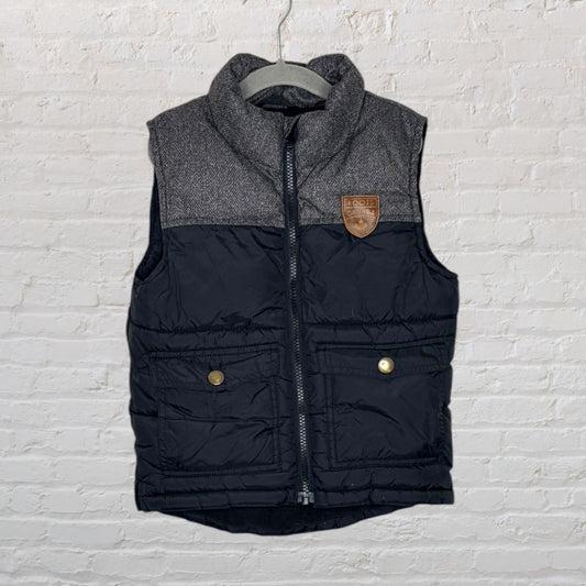 Black and grey quilted vest with tweed accents and a Roots Canada leather patch, featuring front pockets and a zip closure.