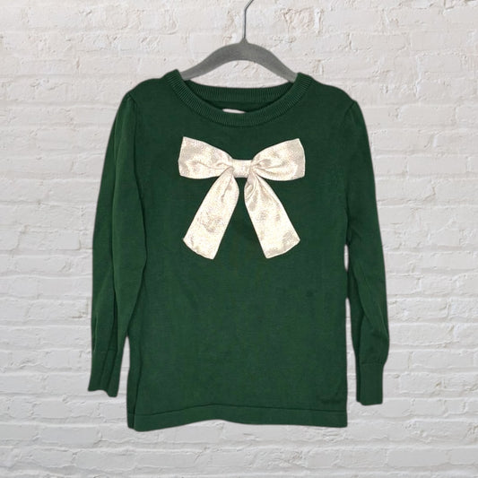 Green sweater with a large decorative beige bow on the front, hanging on a hanger against a white brick background.