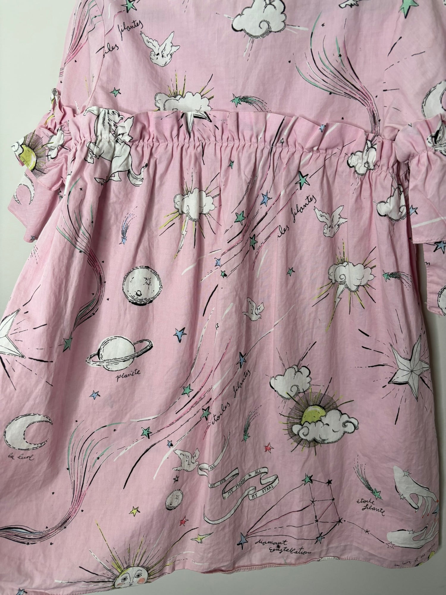 Pink dress with celestial illustrations and ruffled sleeves hanging against a white brick background.
