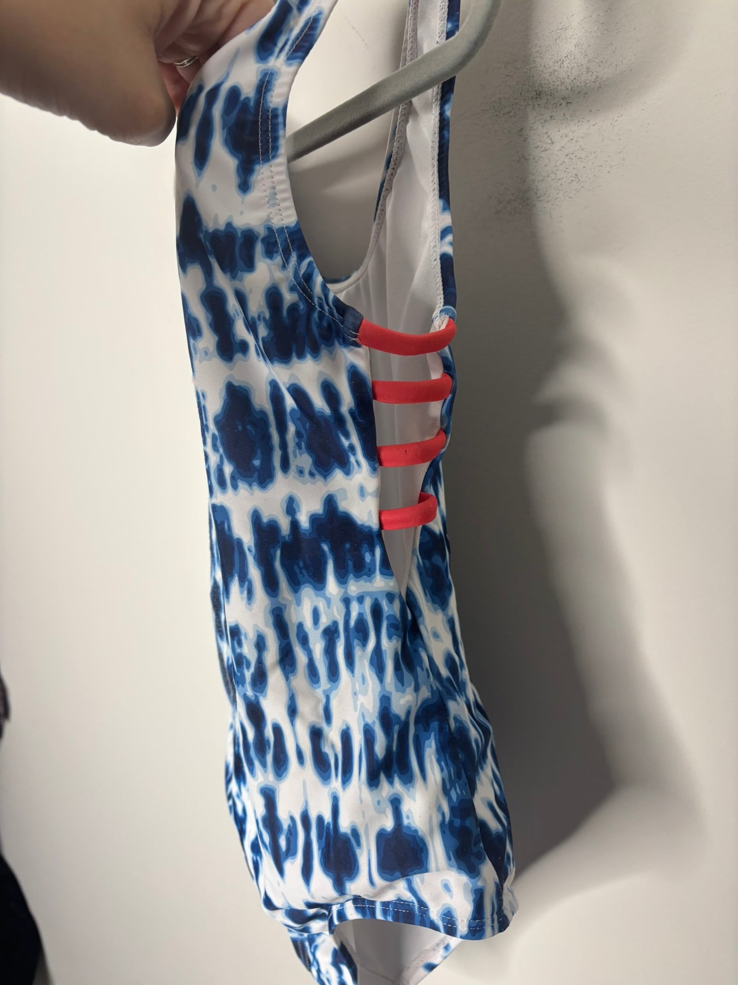 Blue and white tie-dye one-piece swimsuit with cross back detail, displayed on a hanger against a brick wall.