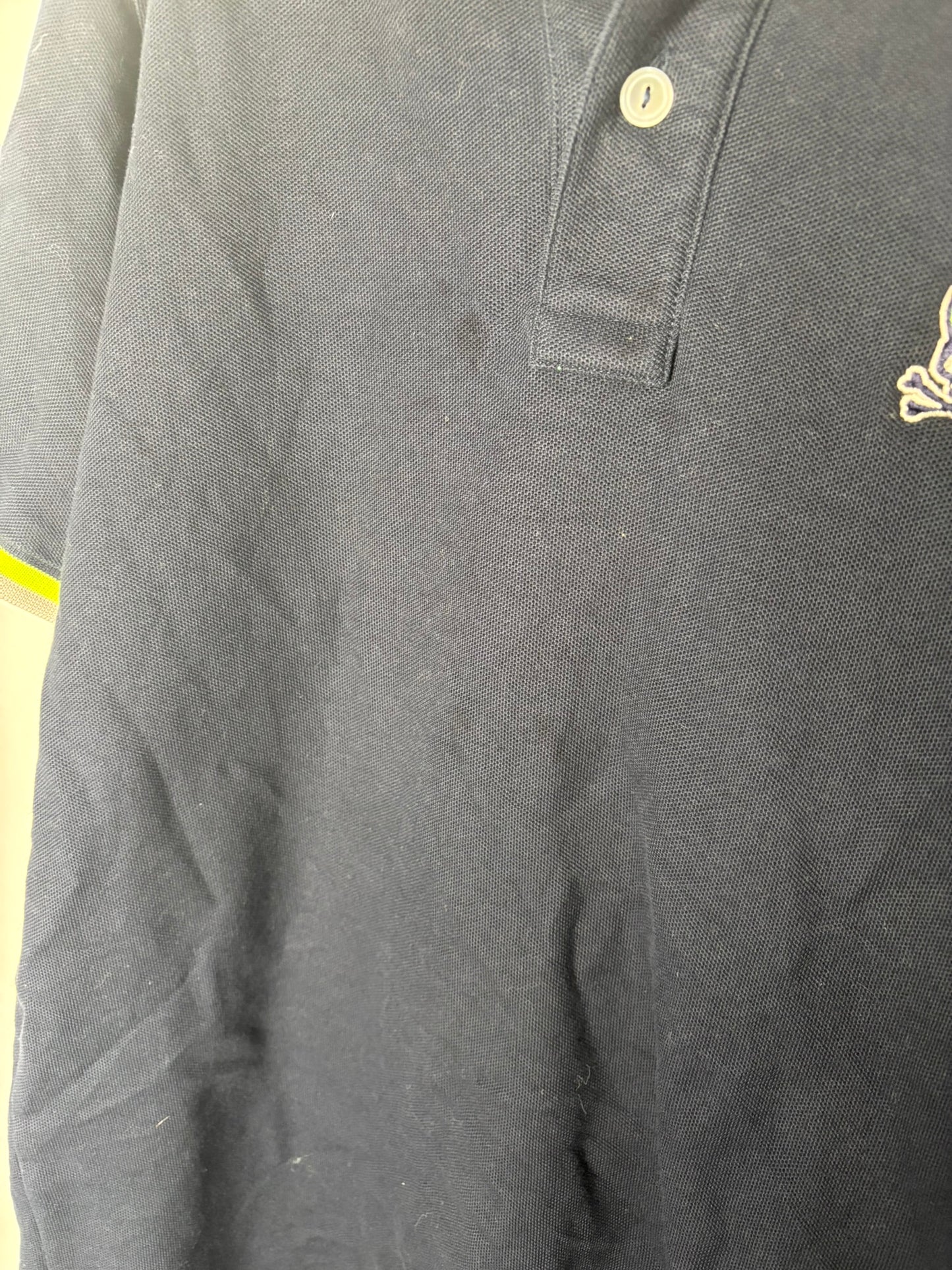 Navy Psycho Bunny polo shirt with neon yellow trim hanging on a grey hanger against a white brick wall.