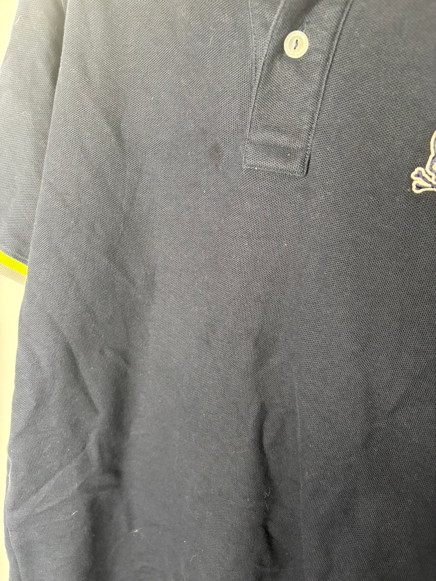 Navy Psycho Bunny polo shirt with neon yellow trim hanging on a grey hanger against a white brick wall.