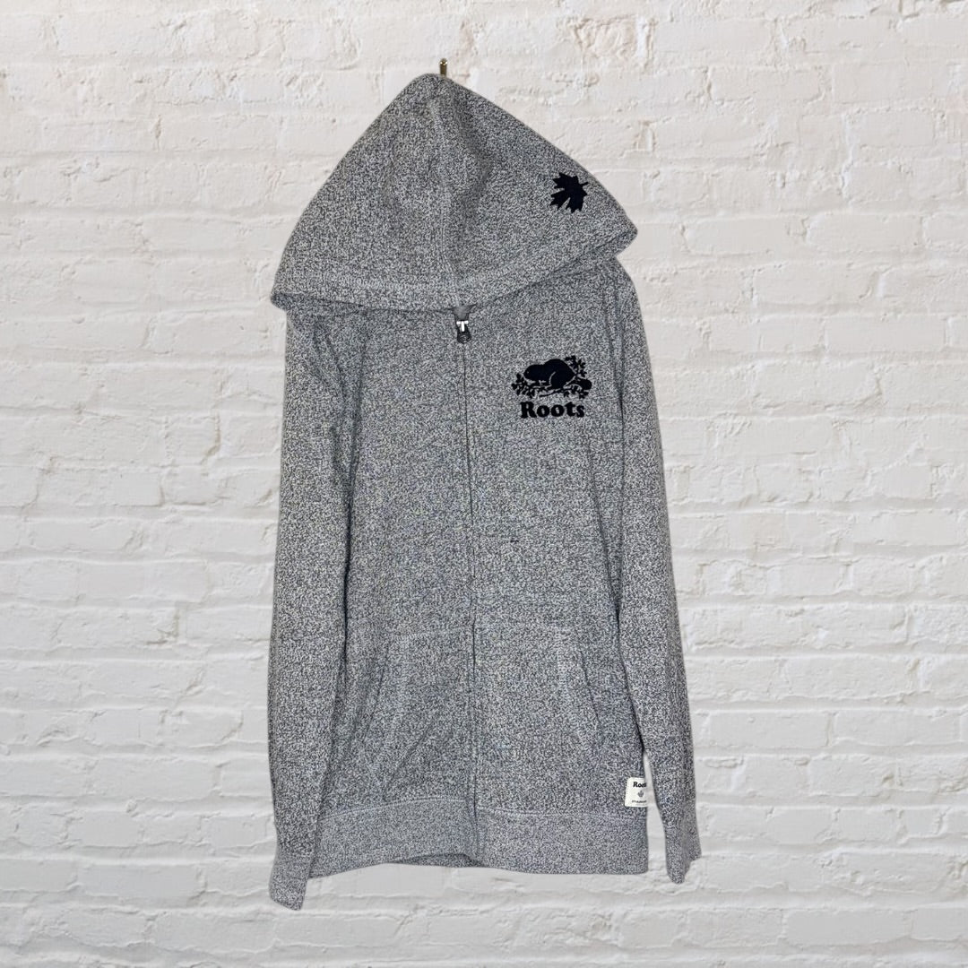 Grey Roots full-zip hooded sweatshirt with logo on chest and maple leaf on hood, hanging against a white brick wall.